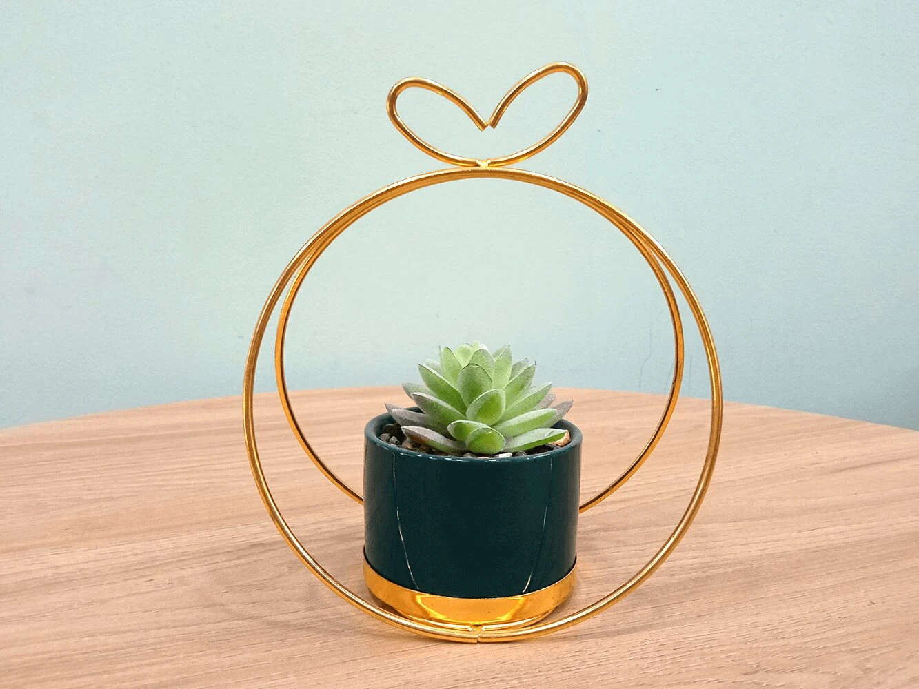 Maceta colgante dorada con forma de círculo y corazón, contiene suculenta artificial verde en maceta verde y dorada.