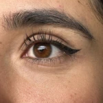 Ojo marrón con delineado cat eye y cejas pobladas, piel con textura visible.