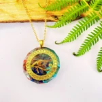 Colgante Orgonita Ojo de Horus dorado con siete minerales y cadena, sobre fondo blanco con helecho y madera.