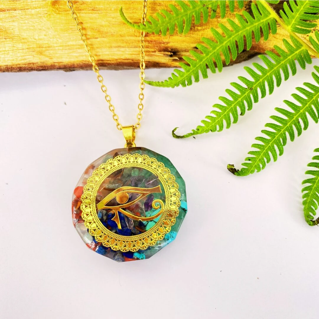 Colgante Orgonita Ojo de Horus dorado con siete minerales y cadena, sobre fondo blanco con helecho y madera.