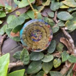 Colgante de orgonita redondo con símbolo Om dorado, incrustaciones de siete cuarzos y detalles florales dorados, entre hojas verdes.