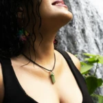 Colgante de piedra aventurina verde con cadena negra, en un primer plano de mujer, con cascada y follaje de fondo.