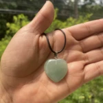 Colgante corazón verde jade con cordón negro sostenido en mano, pulsera con cuentas verdes y doradas al fondo.