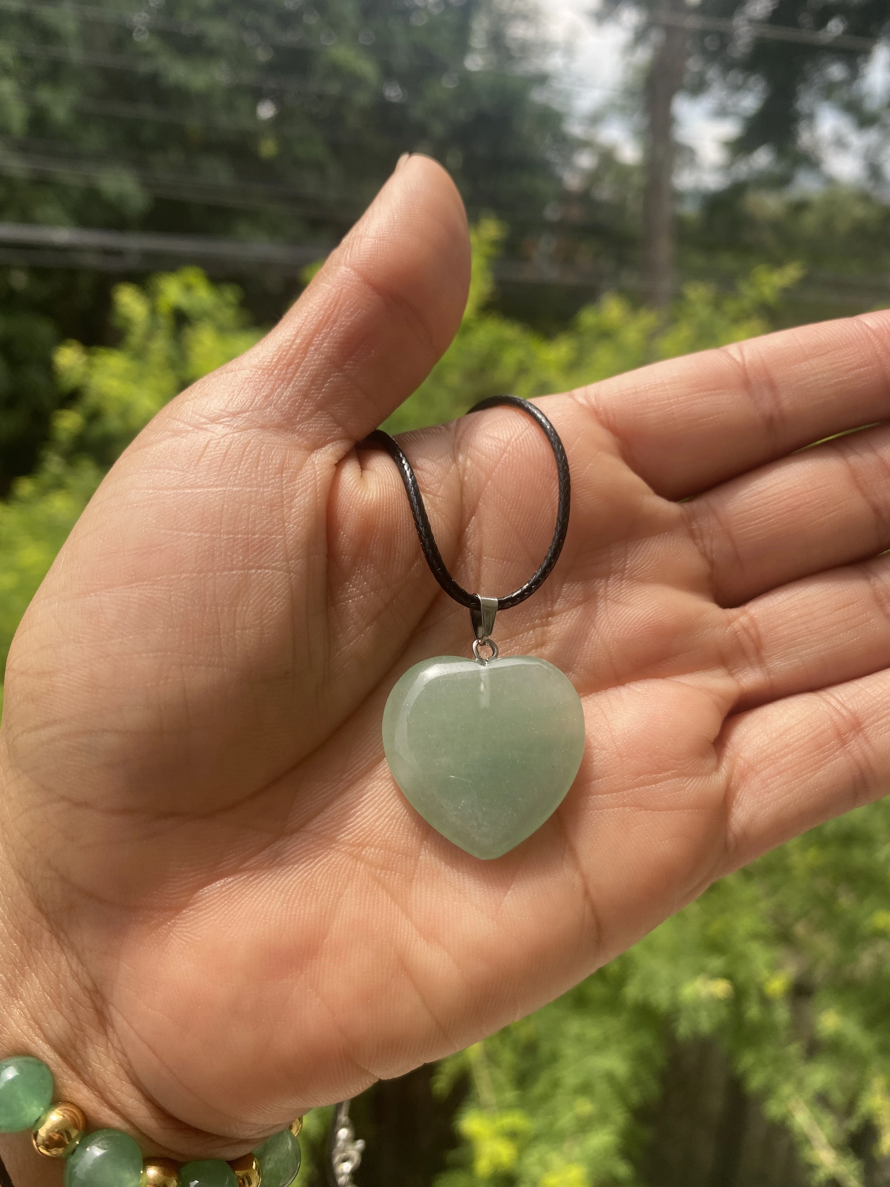 Colgante corazón verde jade con cordón negro sostenido en mano, pulsera con cuentas verdes y doradas al fondo.