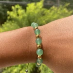 Pulso de persona con pulsera de aventurina verde y cuentas doradas, fondo de follaje verde.