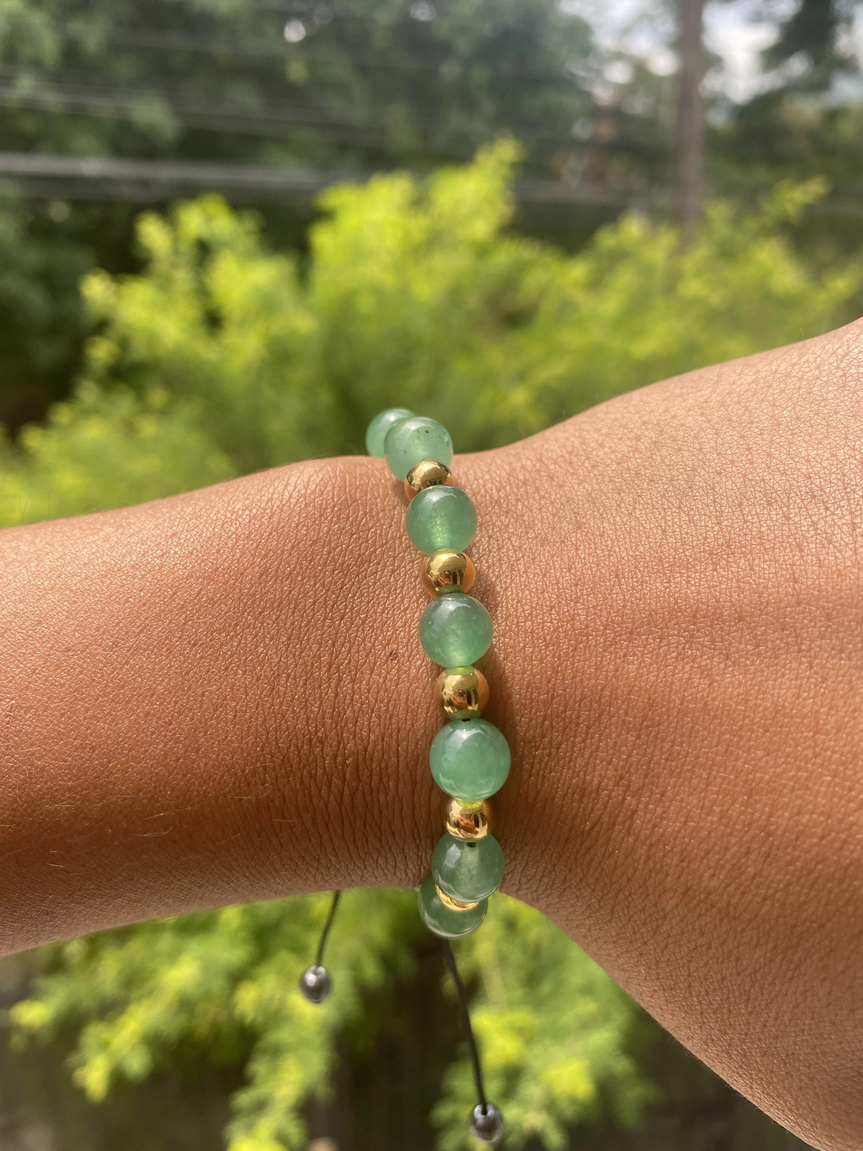 Pulso de persona con pulsera de aventurina verde y cuentas doradas, fondo de follaje verde.