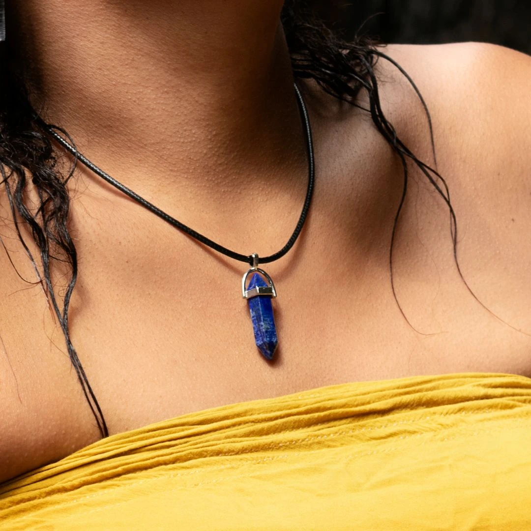 Colgante de lapislázuli azul en forma de cristal facetado, con cadena negra, sobre piel y tela amarilla.