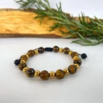 Pulsera ajustable de ojo de tigre y cuentas doradas, sobre fondo blanco y madera, con ramas de romero.