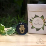 Orgonita redonda azul con mano de Fátima dorada incrustada, al lado de empaque y plantas.