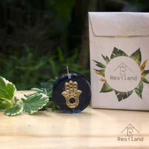 Orgonita redonda azul con mano de Fátima dorada incrustada, al lado de empaque y plantas.