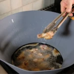 Frywall de silicona color gris instalada en una sartén, protegiendo contra salpicaduras de aceite mientras se fríen piezas de pollo con pinzas.