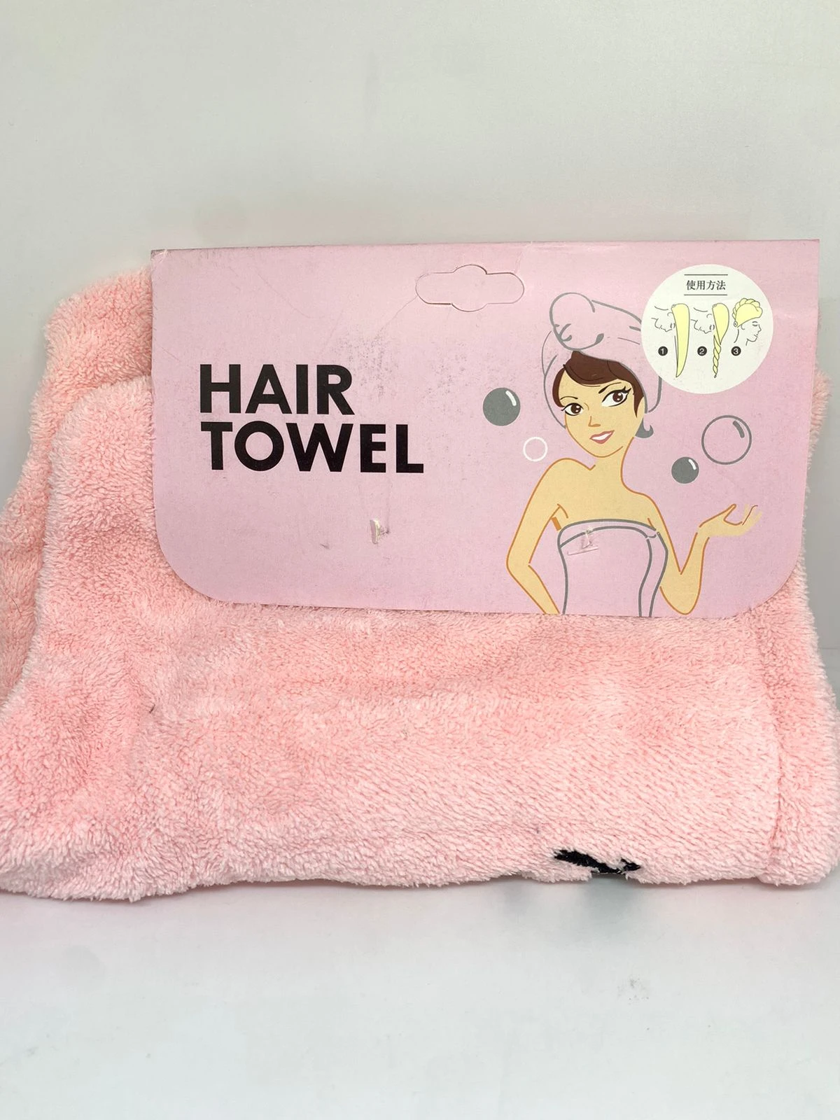 Toalla de microfibra color rosa para el cabello, con empaque que muestra instrucciones gráficas de uso y una de mujer con el producto puesto.