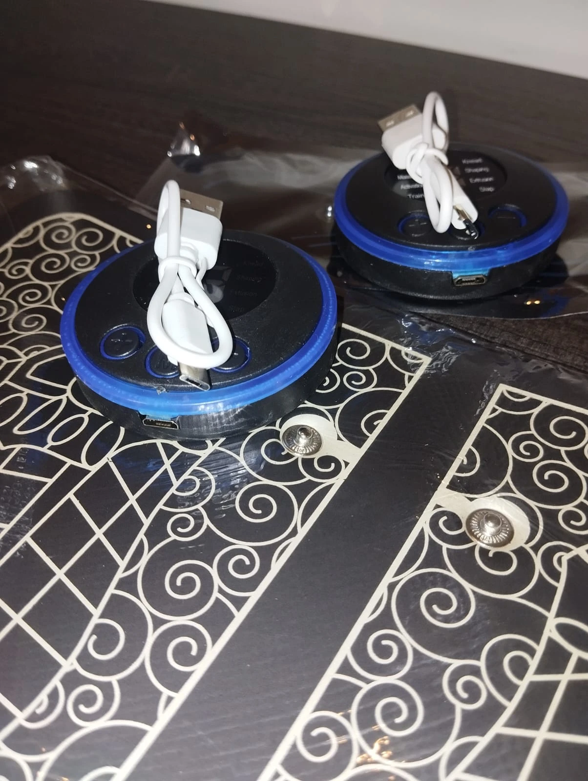 Combo Tapete Masajeador y Mariposa Masaj: dos dispositivos circulares negros con bordes azules y cables blancos USB conectados.