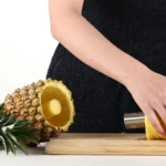 Pelador de piña cortando rodajas de piña amarilla sobre tabla de madera.