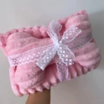 Kit cobija y almohada doble faz rosa, suave, con encaje blanco y moño, sostenido en mano.