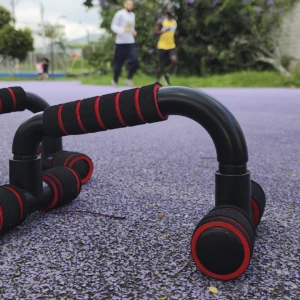 Soportes para flexiones negros con agarres acolchados rojos y base circular en suelo morado. Personas corriendo al fondo.