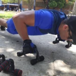 Hombre realizando flexiones con soportes negros y azules de ejercicio en exteriores.