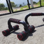 Soportes para flexiones negros con agarres rojos y acolchados, sobre superficie de cemento con valla metálica al fondo.