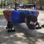 Hombre en flexión con soportes para push up negros, shorts azules. Vista cercana.