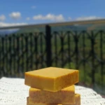 Torre de cuatro jabones artesanales de cúrcuma con motas exfoliantes, sobre mantel de encaje blanco con fondo de paisaje montañoso y cielo despejado.