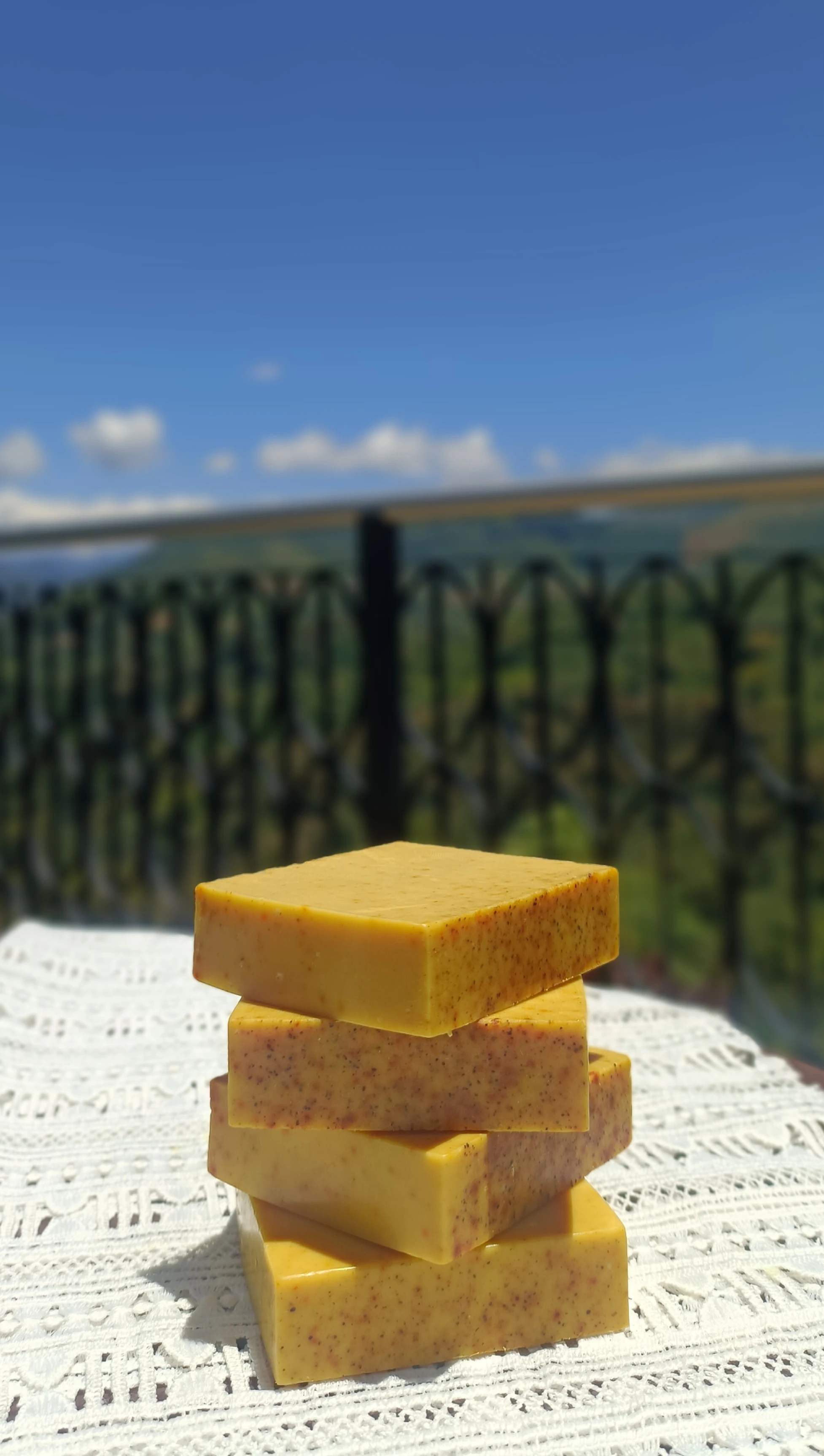 Torre de cuatro jabones artesanales de cúrcuma con motas exfoliantes, sobre mantel de encaje blanco con fondo de paisaje montañoso y cielo despejado.