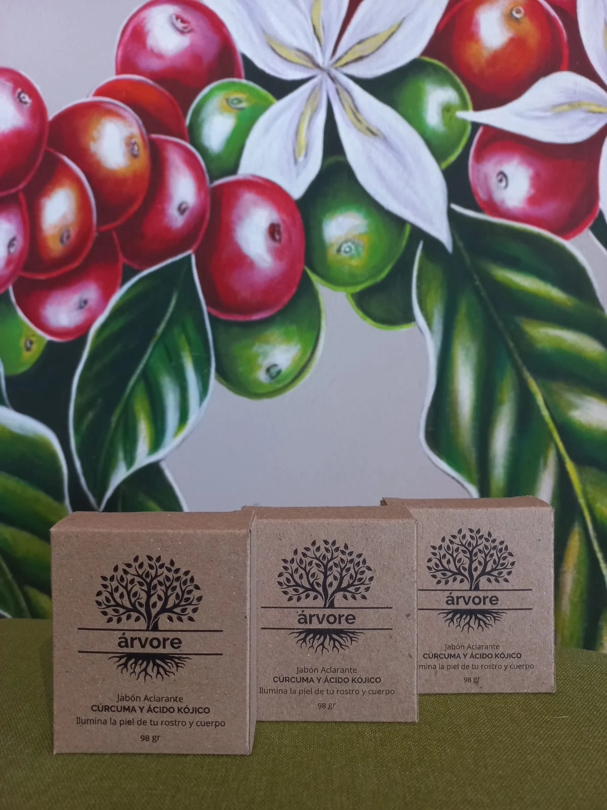 Tres jabones aclarantes Árvore de cúrcuma y ácido kójico, cada uno de 98 gramos, en cajas de cartón sobre fondo con mural de café y flores.