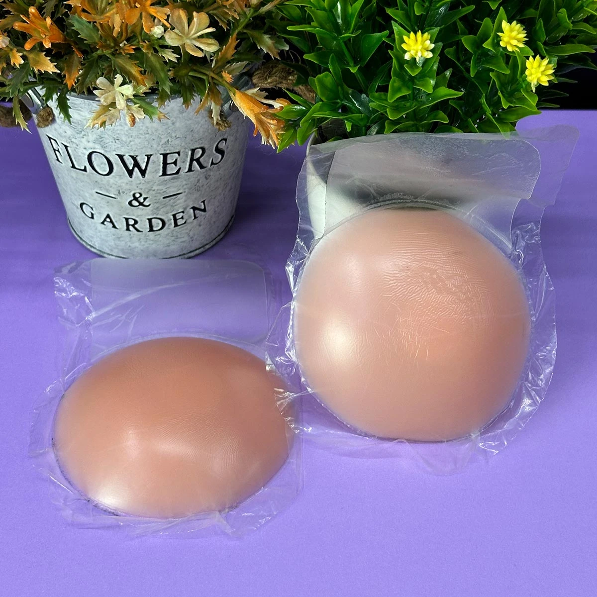 Dos cubrepelzones levantadores de senos en silicona color piel, presentados en empaques individuales transparentes sobre fondo morado junto a materas.