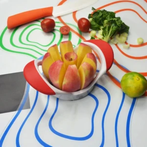 Picador de manzana rojo y blanco cortando una manzana sobre mantel de cocina con frutas y cuchillo.