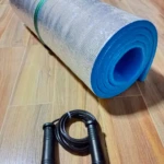 Colchoneta de ejercicio plateada y azul enrollada junto a un lazo negro con mangos acanalados.