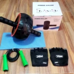 Rueda de abdominales negra con aros rojos, guantes de gimnasio negros y cuerda de saltar verde sobre fondo de madera.