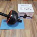 Rueda de ejercicio para abdominales, negra con detalles rojos, sobre tapete azul, junto a caja.
