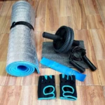 Kit de entrenamiento con rodillo ab, cuerdas, guantes y banda de resistencia azul sobre piso de madera.