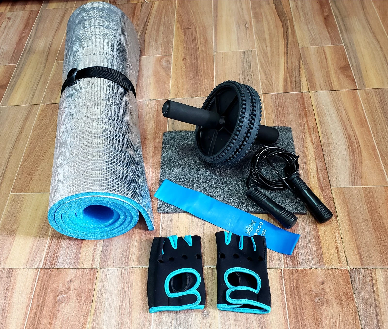 Kit de entrenamiento con rodillo ab, cuerdas, guantes y banda de resistencia azul sobre piso de madera.