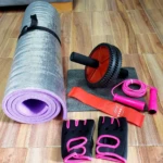 Kit de ejercicio con tapete morado enrollado, rueda abdominal roja, banda elástica naranja, cuerda de saltar rosa y guantes negros con detalles rosas.