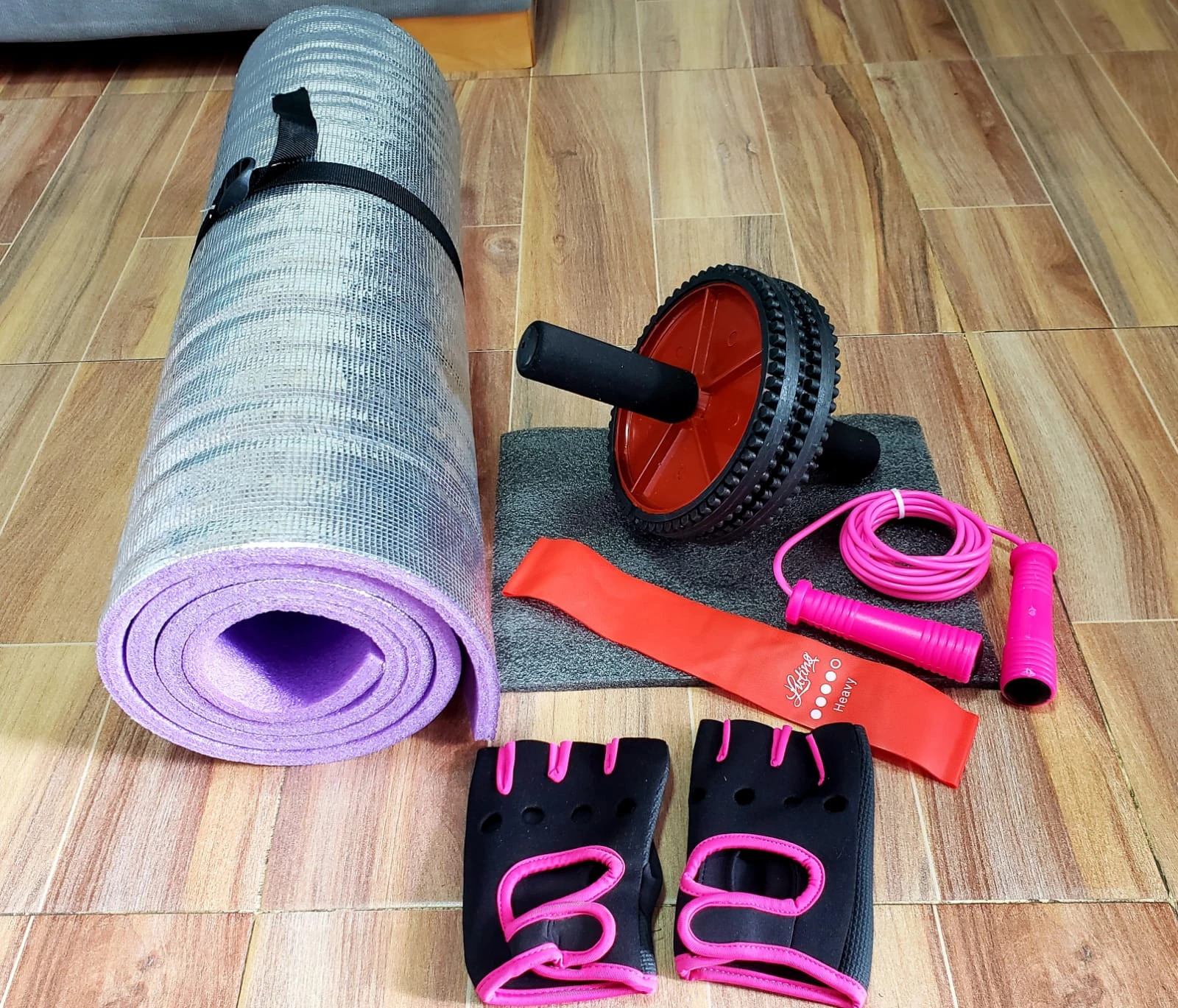 Kit de ejercicio con tapete morado enrollado, rueda abdominal roja, banda elástica naranja, cuerda de saltar rosa y guantes negros con detalles rosas.