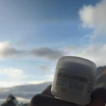 Frasco de 60g de crema aclarante Lotux, presentado en envase blanco con tapa enroscable, sostenido a contraluz frente a un cielo nublado y montañas.