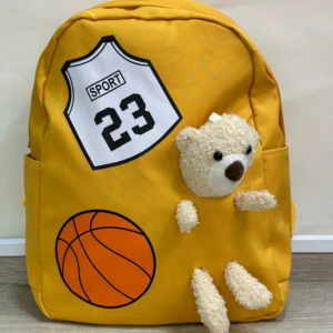 Mochila amarilla escolar con diseño de camiseta deportiva y oso de peluche, ideal para niños.