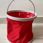 Balde de agua plegable rojo con asa metálica y borde blanco, colocado sobre baldosas.