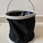 Balde plegable negro de tela con aro blanco y asa metálica, para agua.