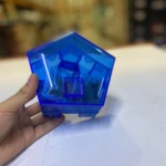 Trampa para cucarachas azul transparente en forma pentagonal, sujeta por mano. Contiene pequeñas bolsas y compartimento central cuadriculado.