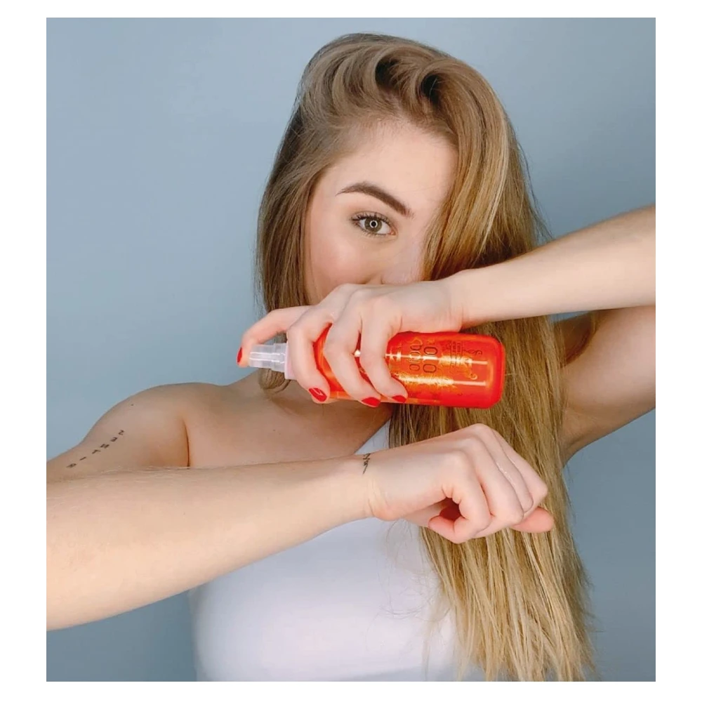 Mujer rubia sosteniendo un frasco naranja de loción aclaradora con manzanilla y pantenol frente a su rostro, mostrando la textura del producto en su mano.