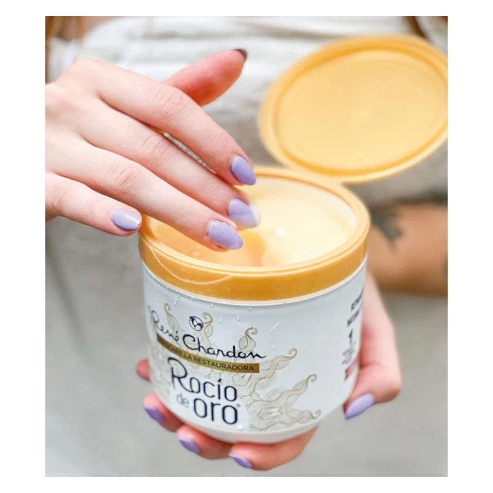 Manos con esmalte lila sostienen un frasco de Mascarilla Restauradora Rocío de Oro, mostrando su textura cremosa y el envase destapado color dorado.