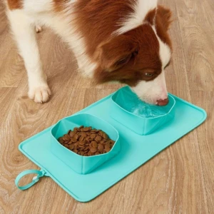 Perro de pelaje café y blanco bebiendo agua de un comedero plegable de silicona color turquesa, que incluye dos recipientes integrados en un tapete.