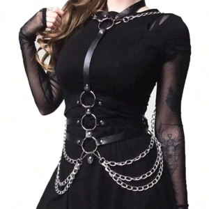 Arnés metal corsé negro con tiras de cuero sintético, aros metálicos verticales al frente y cadenas decorativas colgantes sobre un vestido negro.