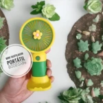 Mini ventilador portátil amarillo y verde con diseño de flor, sostenido por mano sobre fondo de flores y tierra decorativa.