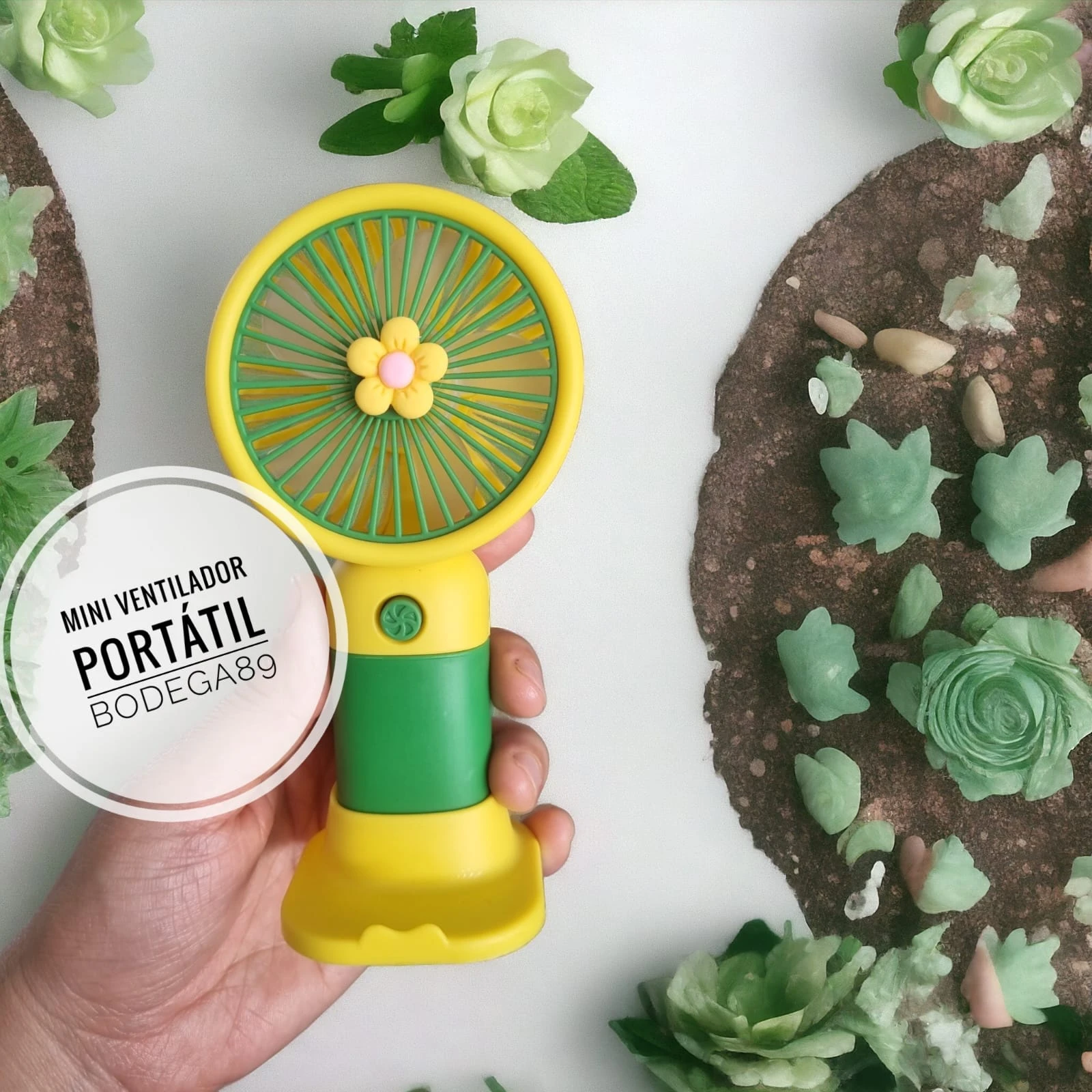 Mini ventilador portátil amarillo y verde con diseño de flor, sostenido por mano sobre fondo de flores y tierra decorativa.