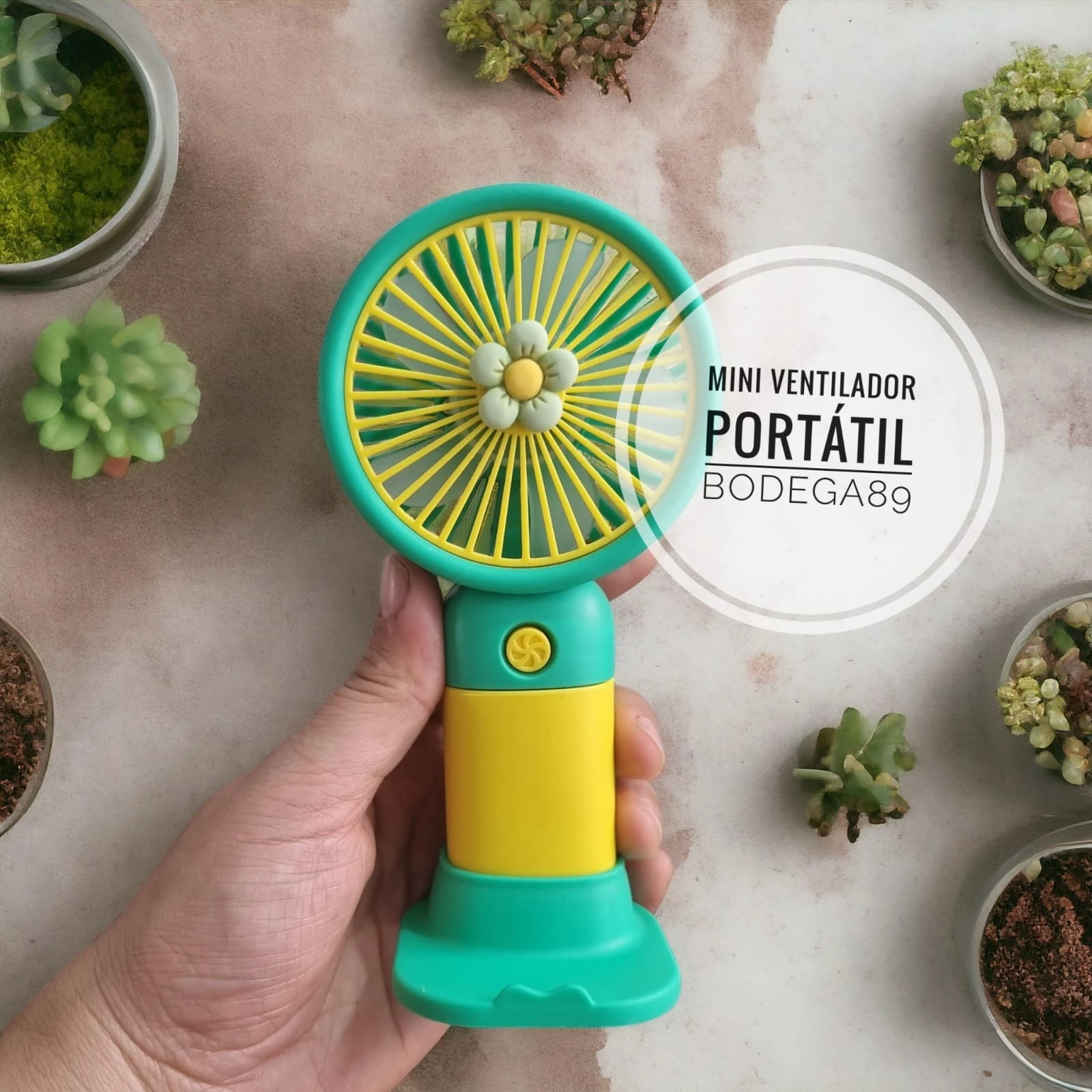 Mini ventilador portátil amarillo y verde con diseño de flor, sostenido por mano, rodeado de suculentas.