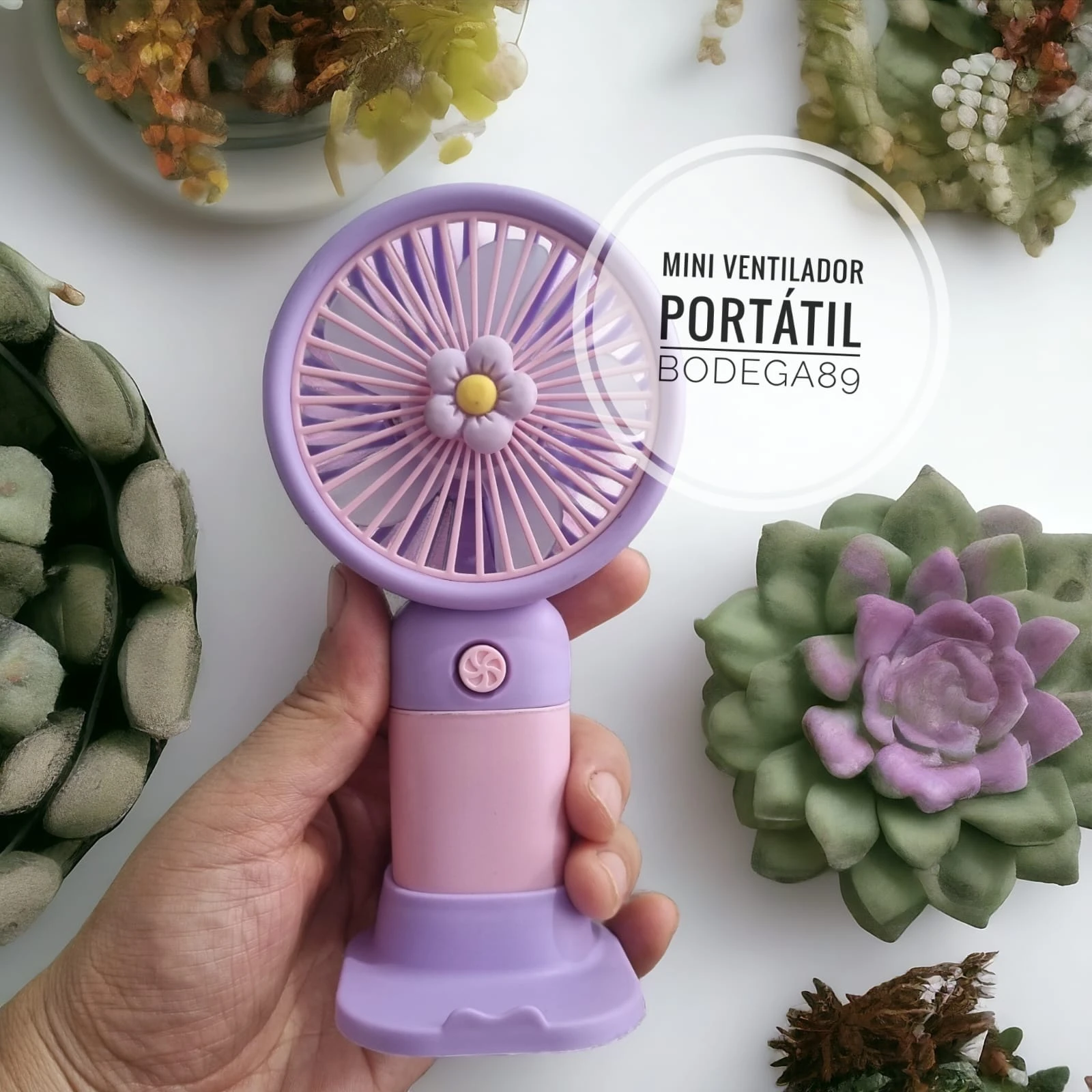 Mini ventilador portátil morado y rosa con diseño de flor, sostenido por mano, al lado de suculentas.