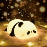 Lámpara de noche táctil en forma de panda acostado, hecha de silicona suave con luz cálida, descansando sobre una tela con luces decorativas de fondo.