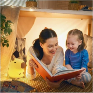 Lámpara infantil de silicona con forma de capibara de luz cálida, ubicada en una acogedora carpa de juegos junto a una madre y su hija leyendo un libro.